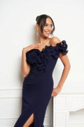 A woman modeling a navy off-the-shoulder evening gown with a ruffled floral bodice and a thigh-high slit, posed against a white paneled wall.