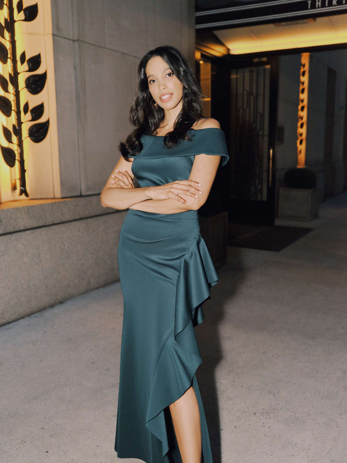 Woman in a teal off-shoulder dress standing in an indoor setting with decorative elements.
