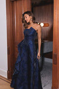 A woman wearing a strapless navy blue evening gown with a textured jacquard pattern and a dramatic layered ruffle skirt, standing in a doorway with a warm light in the background.