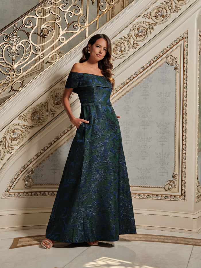 Woman in a dark blue off-shoulder gown standing in an elegant interior setting.