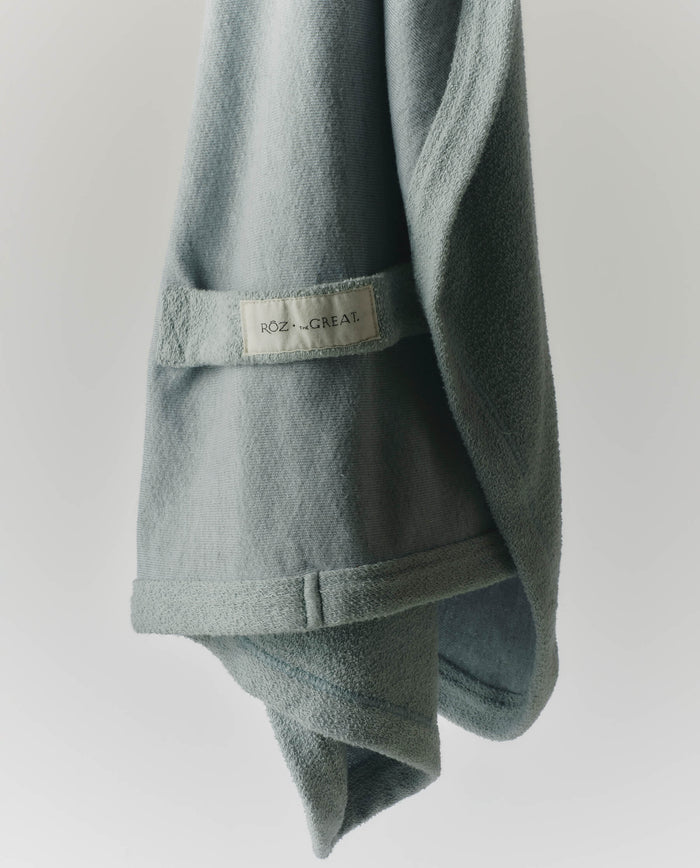 A light green RŌZ + The GREAT The Sweatshirt Hair Towel in Seaglass, from THE GREAT. PF25 ROZ, hangs against a plain white background—ideal for quick drying and reducing frizz.