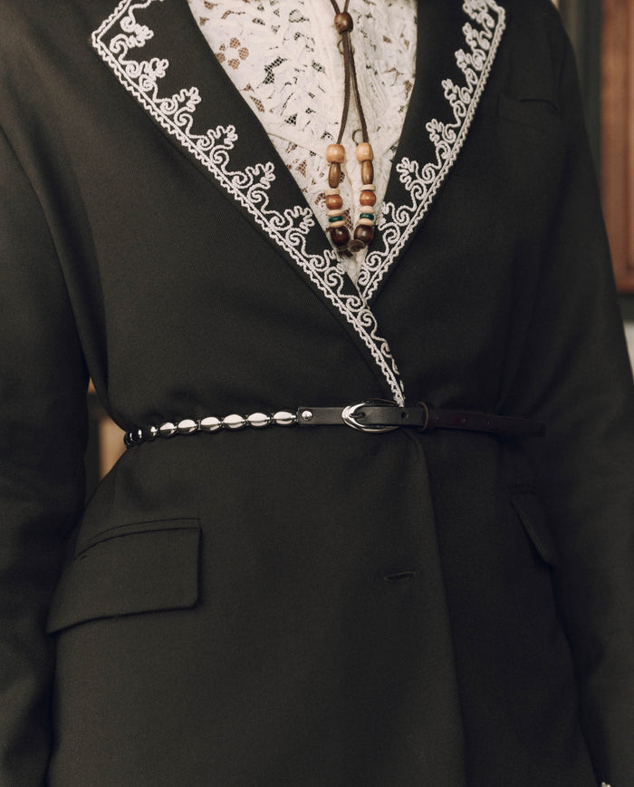 A person wearing a black embroidered jacket with white trim, a white lace shirt, a beaded necklace, and The Beaded Belt in Black With Silver from THE GREAT. F26 STETSON—an elegant nod to classic Western accessories.