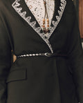 A person wearing a black embroidered jacket with white trim, a white lace shirt, a beaded necklace, and The Beaded Belt in Black With Silver from THE GREAT. F26 STETSON—an elegant nod to classic Western accessories.