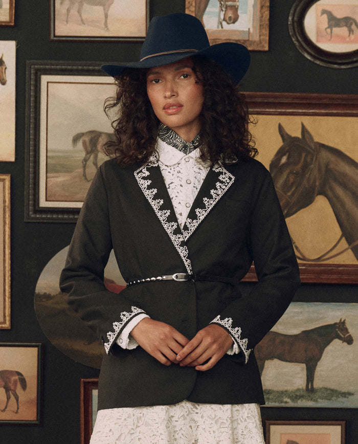 A woman in a western-style outfit, featuring THE GREAT.’s The Beaded Belt in black with silver and a F26 STETSON blue hat, stands before a wall adorned with framed horse paintings.