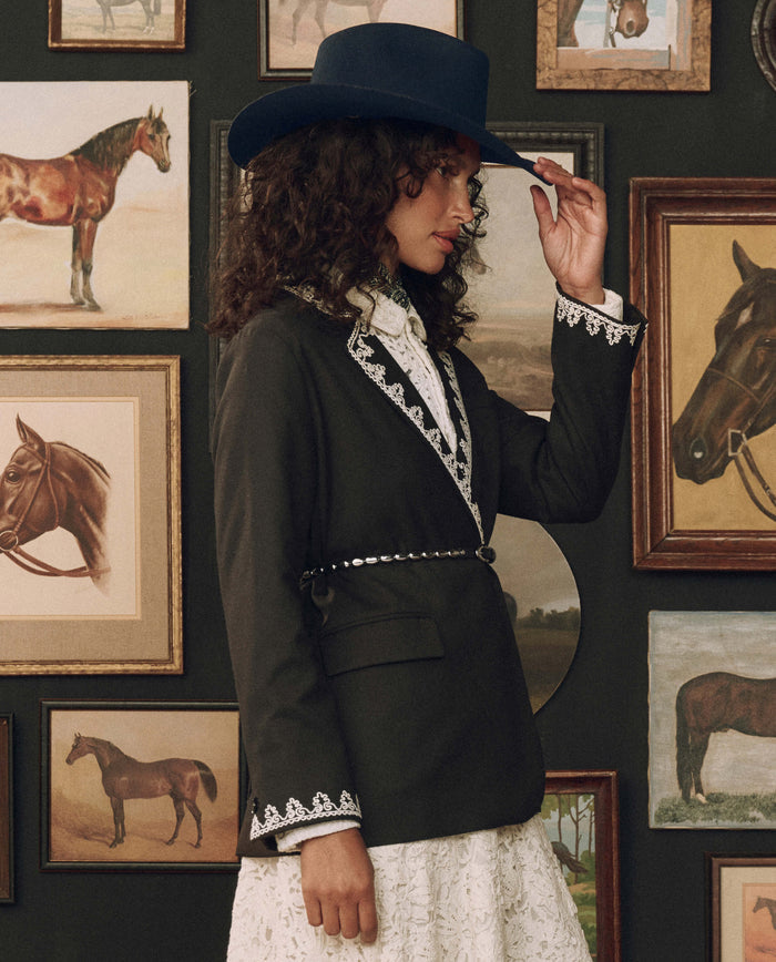 A woman in a dark blazer, white dress, and wide-brimmed hat stands in front of a wall adorned with framed horse paintings, adjusting her hat with one hand. The Beaded Belt in Black With Silver by THE GREAT. F26 STETSON cinches her waist, adding a touch of Western flair to her elegant ensemble.