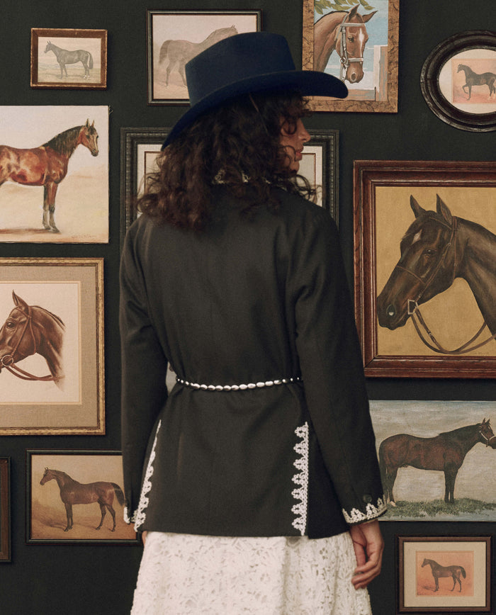 A person in a black jacket and blue hat stands facing a wall adorned with framed horse paintings and drawings, their outfit accented by THE GREAT. F26 STETSON’s The Beaded Belt in Black With Silver—a classic Western accessory.