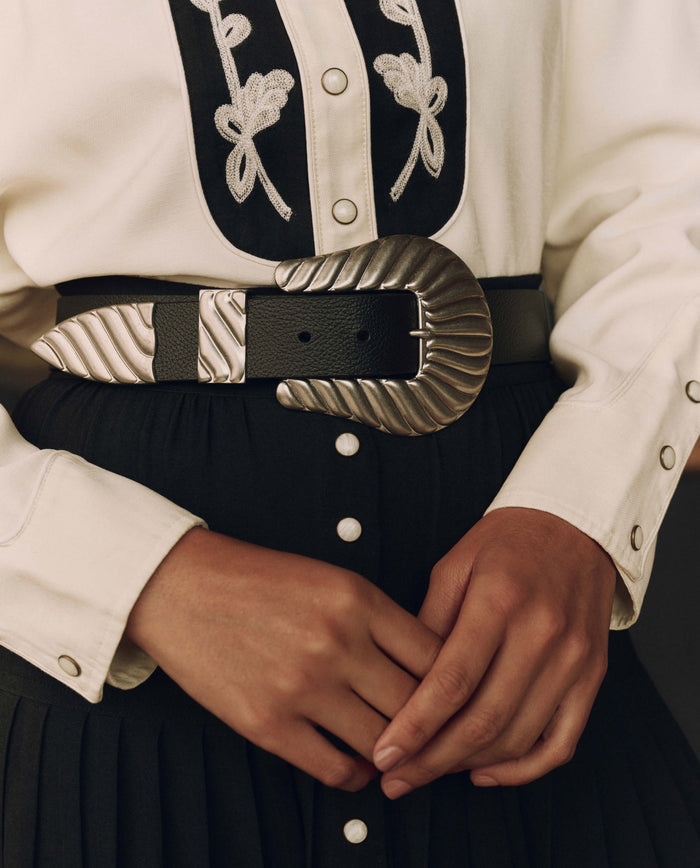 A person wearing a white shirt with embroidered patterns and a black pleated skirt, accessorized at the waist with THE GREAT. F26 STETSON’s Stetson & THE GREAT. The Big Buckle Belt in Black With Silver—a wide top-grain leather belt featuring a large, decorative silver buckle that highlights western craftsmanship. Hands are clasped at the waist.