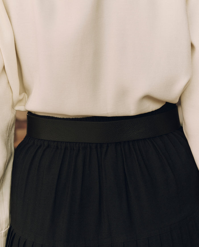 Close-up of a person wearing a cream long-sleeve blouse tucked into a black pleated skirt, accented at the waist with THE GREAT. F26 STETSON Stetson & THE GREAT. The Big Buckle Belt in Black With Silver, showcasing top-grain leather and exceptional western craftsmanship.