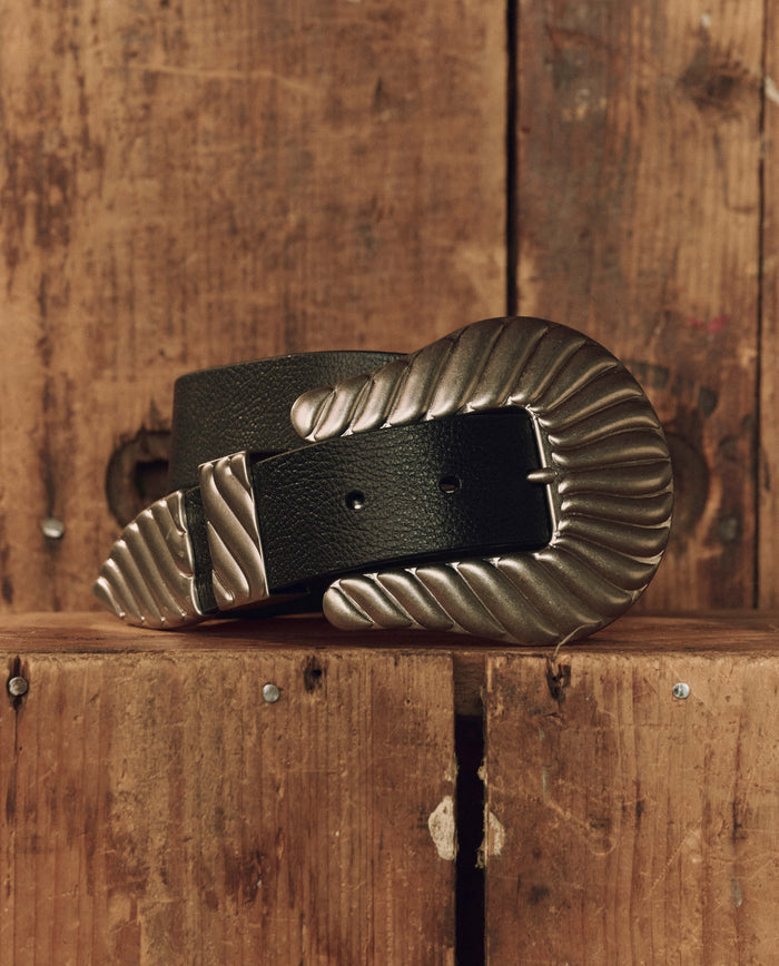 The Stetson & THE GREAT. The Big Buckle Belt in black with silver, from THE GREAT. F26 STETSON, is shown rolled up on a rustic wooden surface, featuring top-grain leather and a textured silver buckle and tip that highlight its timeless western craftsmanship.