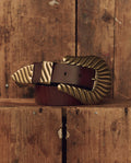 A cognac top-grain leather Stetson & THE GREAT. The Big Buckle Belt—featuring a large, textured brass buckle and tip from THE GREAT. F26 STETSON—is coiled on a wooden surface.