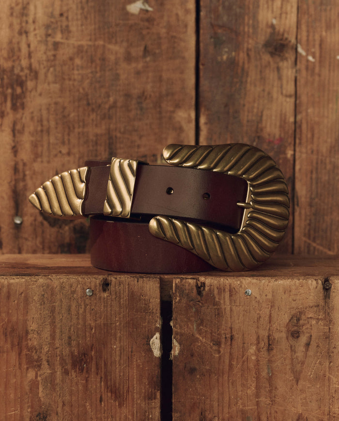 A cognac top-grain leather Stetson & THE GREAT. The Big Buckle Belt—featuring a large, textured brass buckle and tip from THE GREAT. F26 STETSON—is coiled on a wooden surface.
