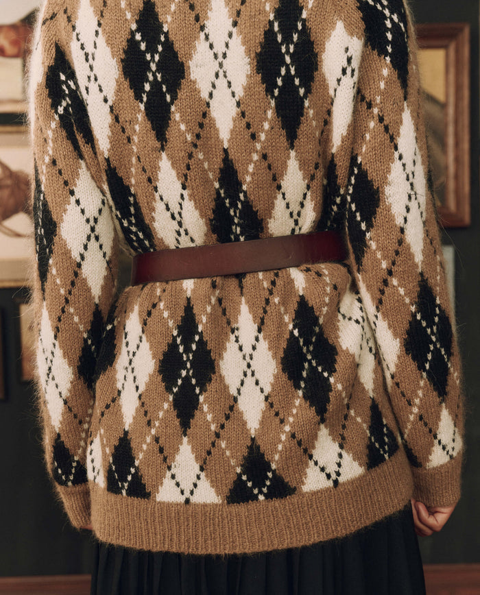 A person is seen from behind wearing a brown, black, and white argyle sweater, styled with THE GREAT. F26 STETSON x Stetson & THE GREAT. The Big Buckle Belt in Cognac With Brass, featuring an oversized buckle, set against a backdrop of framed pictures.