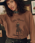 A woman wears THE GREAT. F26 STETSON’s The College Sweatshirt w/ Stetson Graphic in Tobacco, featuring classic “The Great” lettering and a cowboy-on-horse illustration, styled with a black patterned bandana and a hat for an effortlessly cool look inspired by iconic Stetson collaborations.