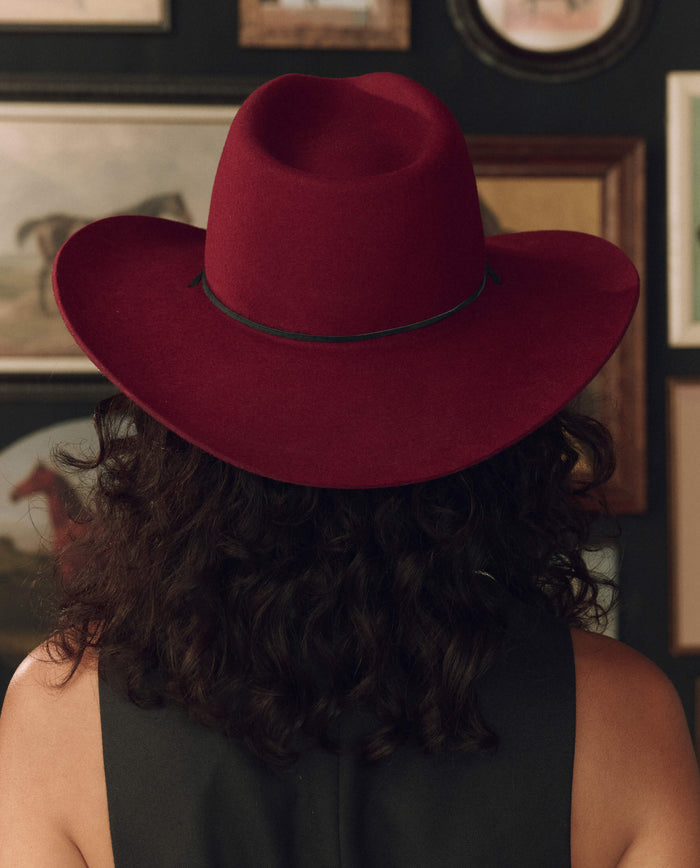 A person with curly dark hair, shown from behind and wearing a black top, models THE GREAT. F26 STETSON’s limited-edition The Ridge Hat in Maroon; framed pictures decorate the wall in the background.