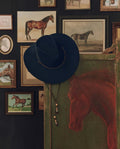 A navy The Ridge Hat by THE GREAT. F26 STETSON hangs on a green panel painted with a horse, with several framed horse paintings and drawings displayed on the wall behind.