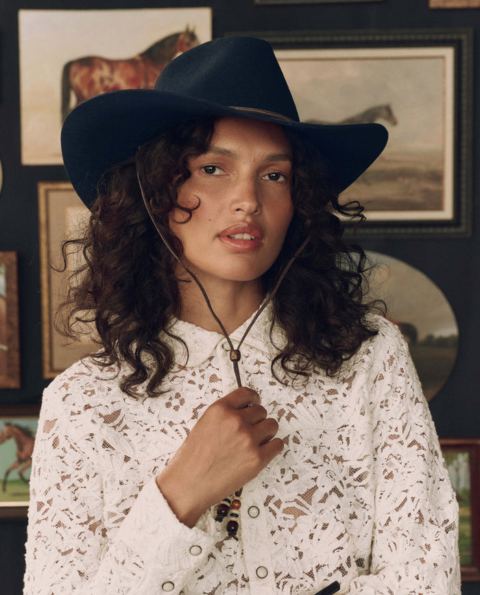 A woman with curly hair wears The Ridge Hat in navy by THE GREAT. F26 STETSON and a white lace shirt, standing before a wall adorned with horse paintings.