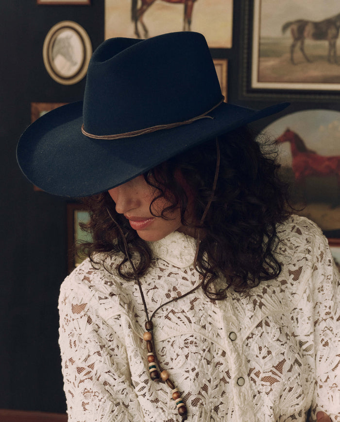 Wearing THE GREAT. F26 STETSON The Ridge Hat in Navy and a white lace shirt, the person looks down with framed horse artwork displayed on the wall behind them.