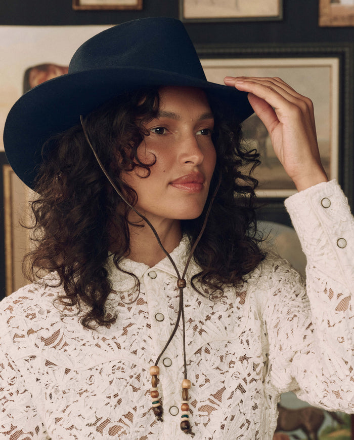 A woman with curly hair, wearing The Ridge Hat in Navy by THE GREAT. F26 STETSON and a white lace shirt, adjusts her hat with one hand while standing indoors in front of framed pictures.