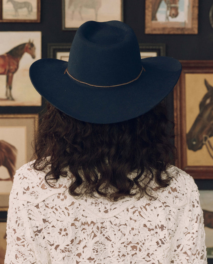 Wearing THE GREAT. F26 STETSON’s The Ridge Hat in navy and a white lace top, a person with curly hair stands facing away before a wall decorated with framed horse artwork.