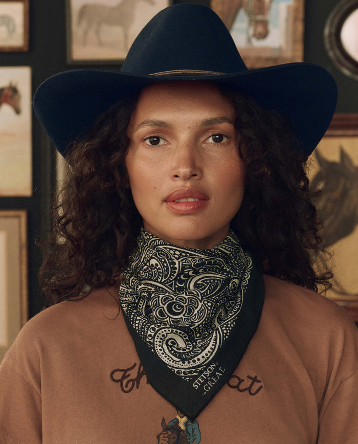 A person stands indoors with framed horse artwork in the background, wearing a dark blue cowboy hat from the Stetson collaboration, a tan shirt, and The Stetson Western Paisley Scarf in black by THE GREAT. F26 STETSON.