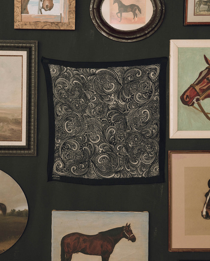 The Stetson Western Paisley Scarf in black by THE GREAT. F26 STETSON, featuring a classic black and white paisley pattern reminiscent of a vintage bandana, is displayed on a dark wall, surrounded by framed paintings and horse illustrations.