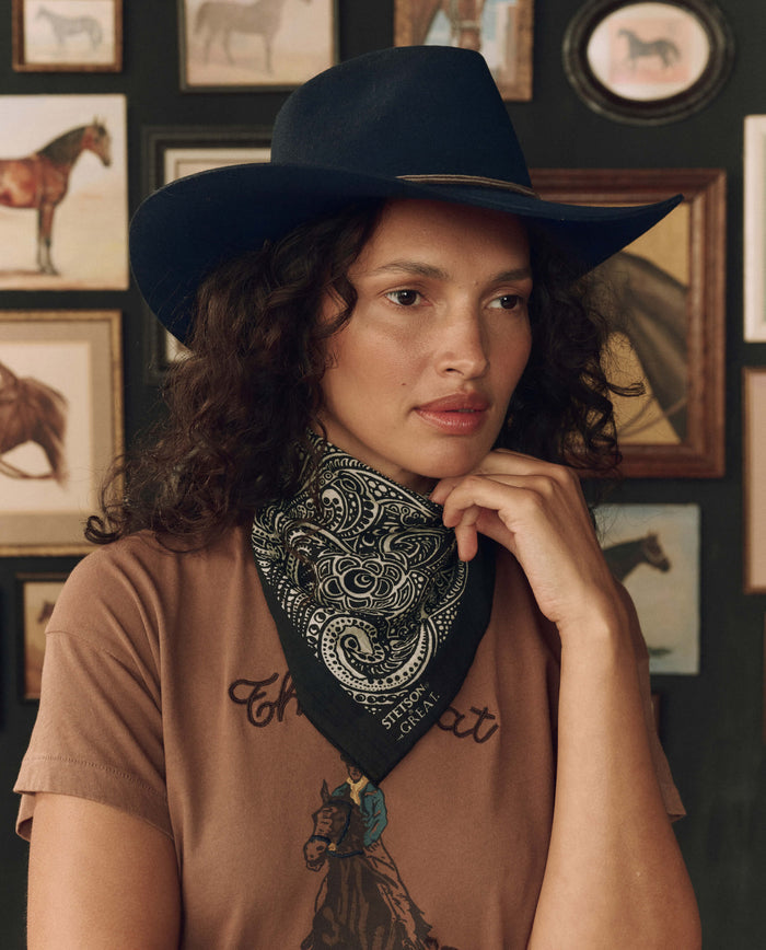 A woman in a cowboy hat and the black Stetson Western Paisley Scarf by THE GREAT. F26 STETSON poses indoors against a wall decorated with framed horse pictures.