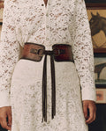 Person wearing a white lace button-up dress with THE GREAT. F26 STETSON’s The Tooled Corset Belt in Burnished Brown, standing in front of framed horse artwork.