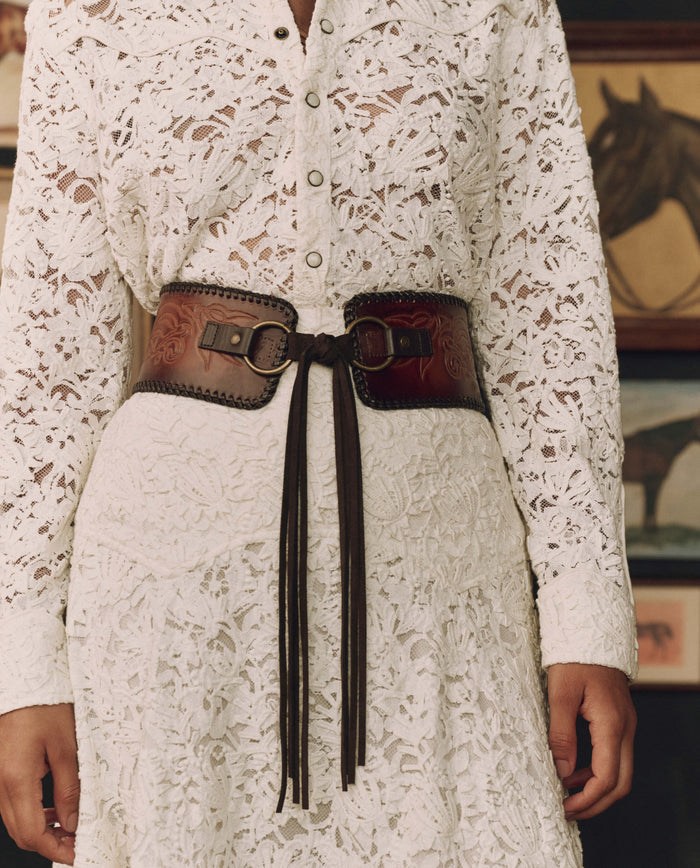 Person wearing a white lace button-up dress with THE GREAT. F26 STETSON’s The Tooled Corset Belt in Burnished Brown, standing in front of framed horse artwork.