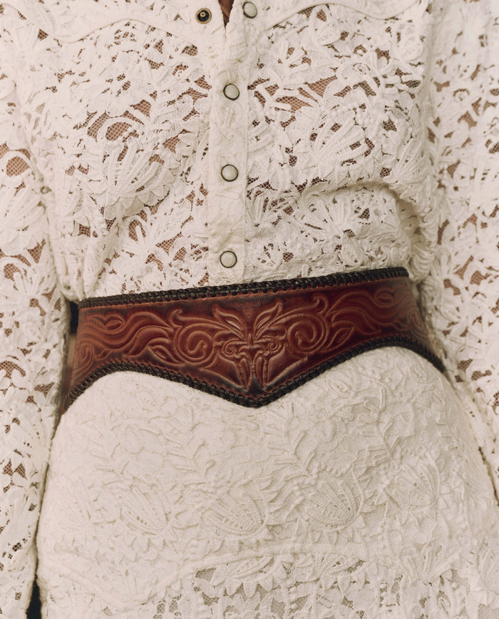 Close-up of a person wearing a white lace buttoned top and skirt, accessorized at the waist with THE GREAT. F26 STETSON’s The Tooled Corset Belt in Burnished Brown.