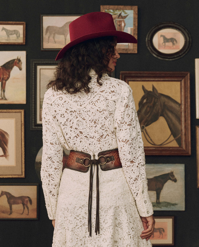 Clad in a white lace dress and a red hat, a person stands with their back to the camera, their silhouette highlighted by The Tooled Corset Belt in Burnished Brown from THE GREAT. F26 STETSON, before a wall adorned with framed horse paintings.
