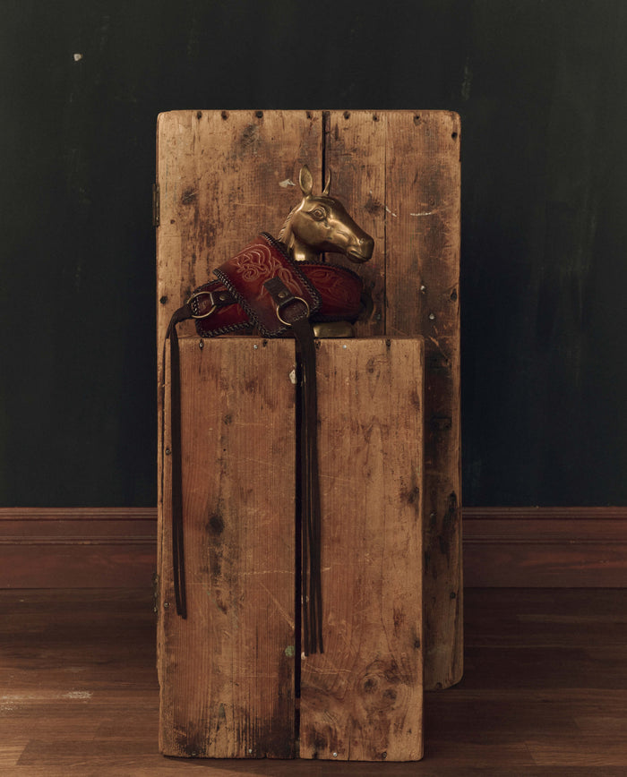 A burnished brown Tooled Corset Belt by THE GREAT. F26 STETSON rests atop a tall, weathered wooden box against a dark wall and wooden floor.