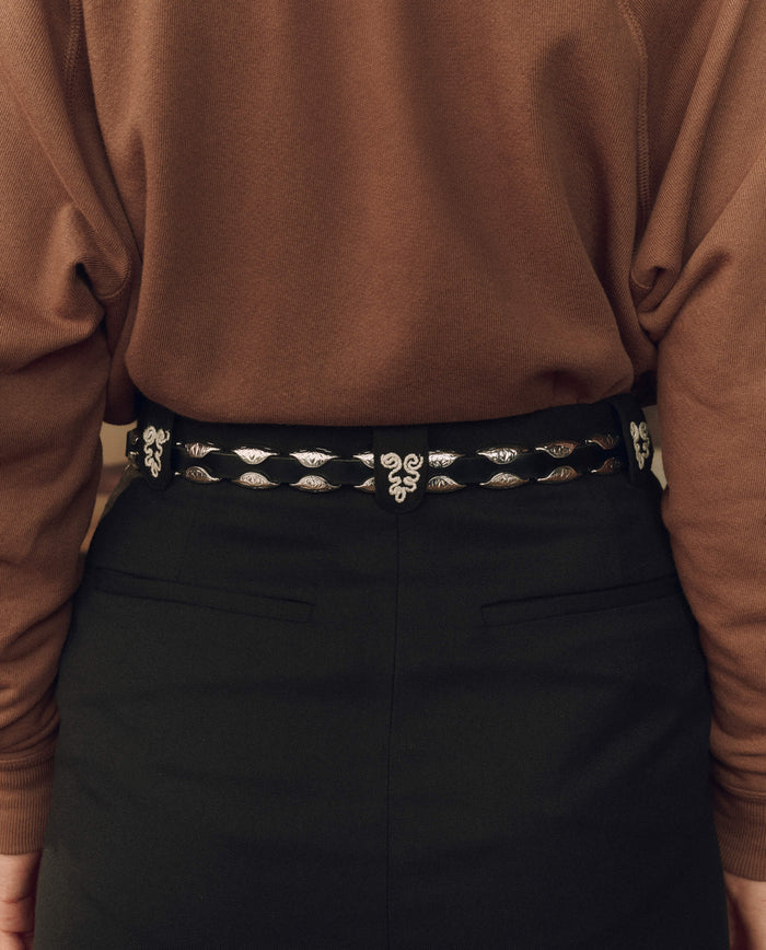 Person wearing a brown long-sleeve top, black high-waisted pants, and THE GREAT. F26 STETSON The Western Hardware Belt in Black with Silver, featuring silver decorative details and a silver-tone buckle, shown from the back.