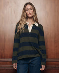 A woman with long wavy hair, wearing THE GREAT. F26 COLLECTORS’ The Academy Polo Sweater in Army League Stripe—a dark green and navy striped rugby sweater with a white collar—paired with jeans, stands in front of a wooden wall.