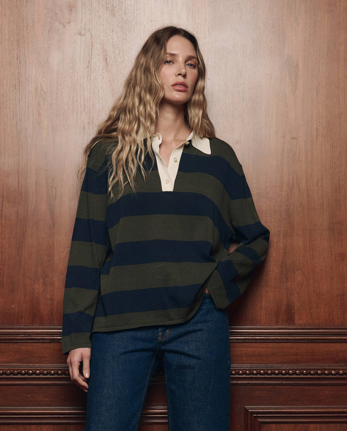 A woman with long, wavy hair stands against a wooden wall, wearing THE GREAT. F26 COLLECTORS The Academy Polo Sweater in Army League Stripe and blue jeans.