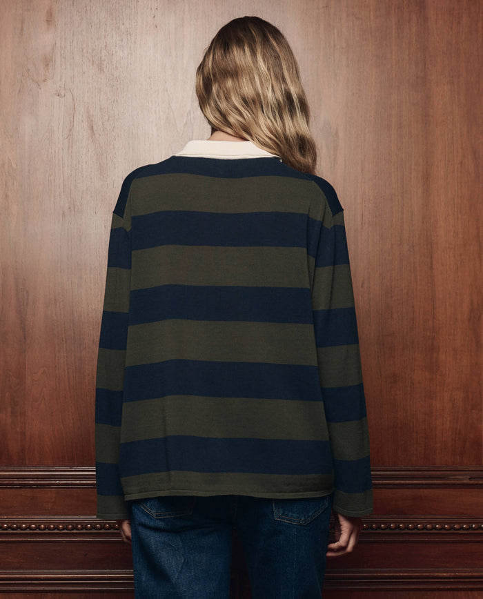 A person with long, wavy hair stands facing a wooden wall, wearing THE GREAT. F26 COLLECTORS The Academy Polo Sweater -- Army League Stripe and blue jeans.