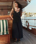 A woman stands on a wooden boat deck by water and palm trees, wearing THE GREAT. SU26's The Apron Dress in black, paired with a light pink neck scarf and brown shoes.