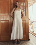 A woman stands on a wooden deck in front of a door, wearing THE GREAT. SU26 The Apron Dress -- Natural with brown boat shoes.