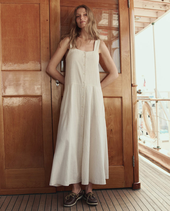 A woman stands on a wooden deck in front of a door, wearing THE GREAT. SU26 The Apron Dress -- Natural with brown boat shoes.