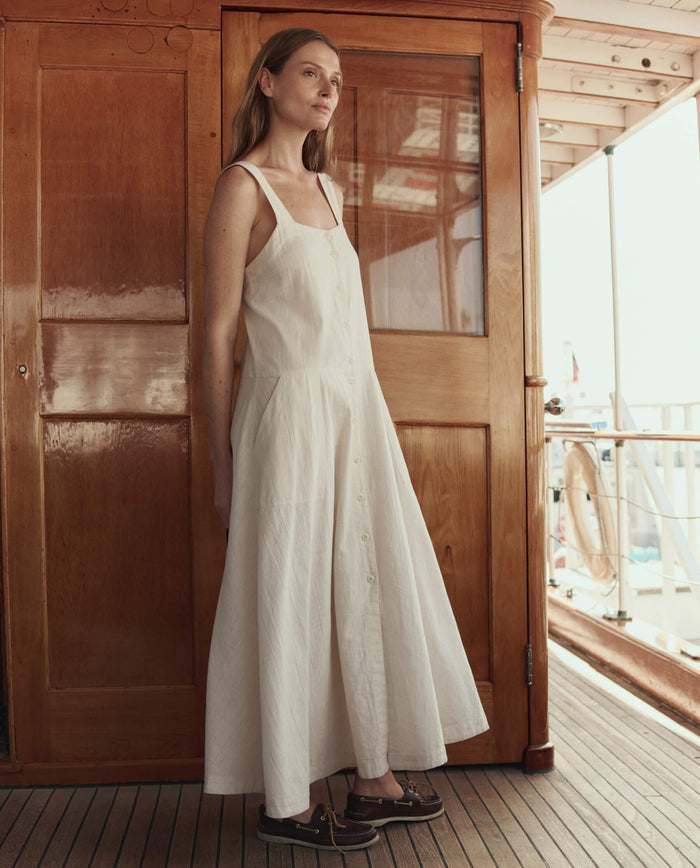 A woman stands on a wooden deck in front of a door, wearing THE GREAT. SU26 The Apron Dress in Natural and dark boat shoes.