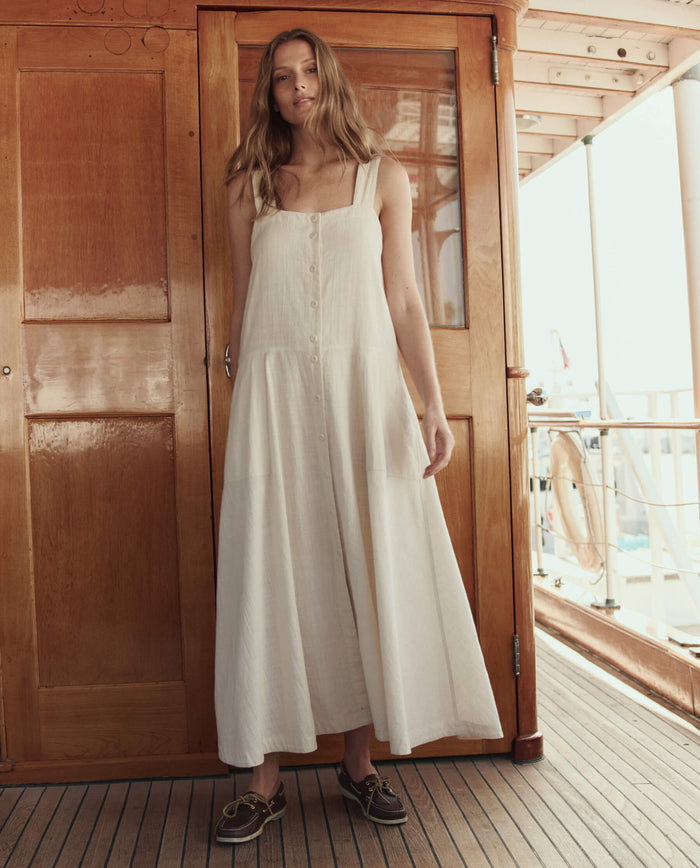 A woman stands on a boat deck in front of wooden doors, wearing THE GREAT. SU26 The Apron Dress. -- Natural, a sleeveless, button-down white dress, paired with dark boat shoes.