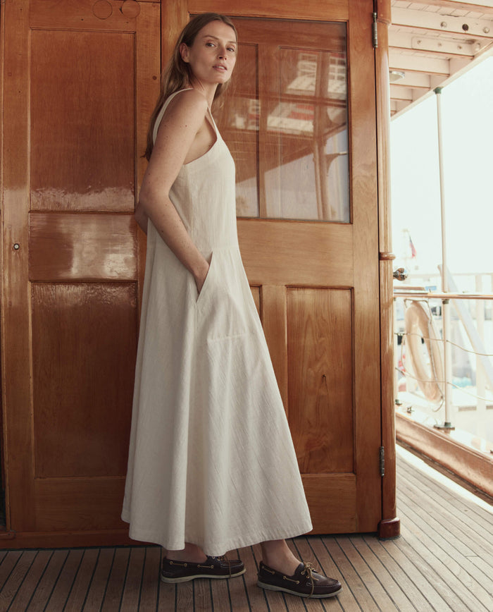 A woman wears THE GREAT. SU26’s The Apron Dress – Natural, a sleeveless long white dress, with dark loafers as she stands on a wooden deck in front of a door, looking toward the camera.