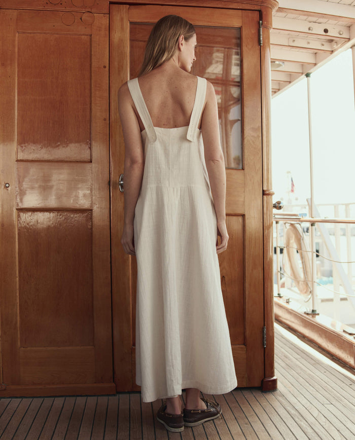 A woman with long hair stands on a wooden deck facing away, wearing THE GREAT. SU26 The Apron Dress in Natural and brown sandals.
