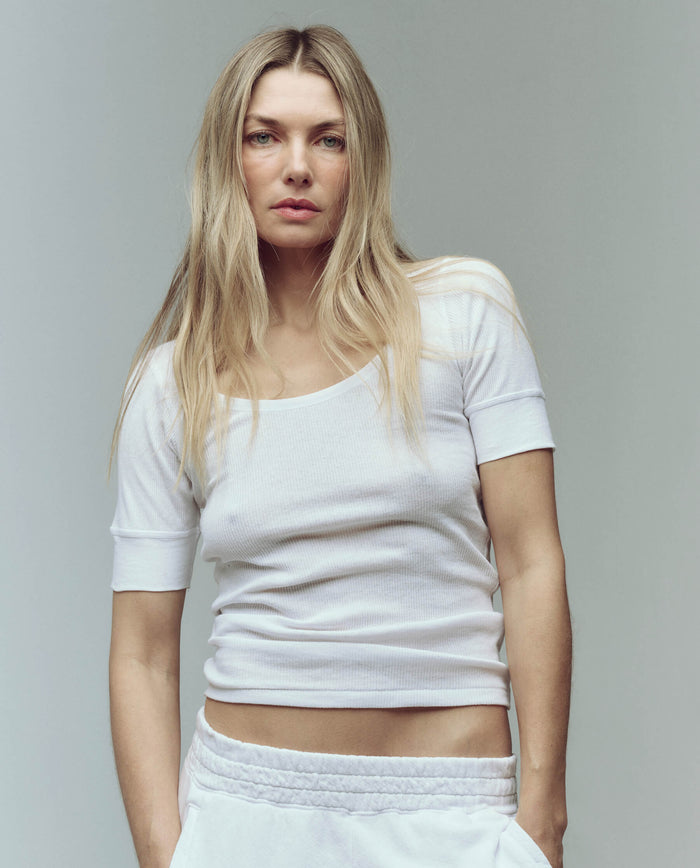 A woman with long blonde hair wears THE GREAT. SU25 BABY RIB The Baby Rib Ballet Tee in True White and matching pants, standing against a plain light background, looking directly at the camera.