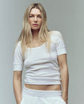 A woman with long blonde hair wears THE GREAT. SU25 BABY RIB The Baby Rib Ballet Tee in True White and matching pants, standing against a plain light background, looking directly at the camera.