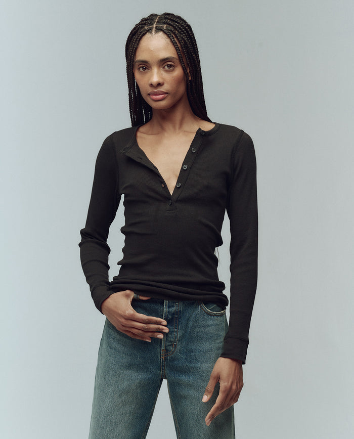 Someone with long braided hair wears THE GREAT. F25 BABY RIB's The Baby Rib Henley in Almost Black, paired with blue jeans, standing against a plain light background.
