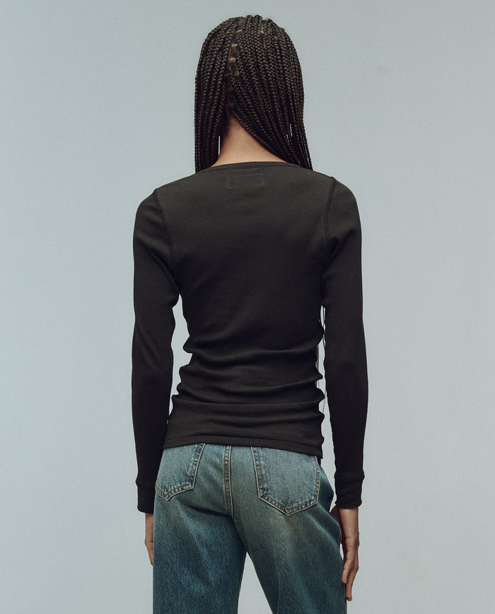 A person with braided hair, wearing THE GREAT. F25 BABY RIB The Baby Rib Henley in Almost Black and blue jeans, stands facing away from the camera against a plain light background.