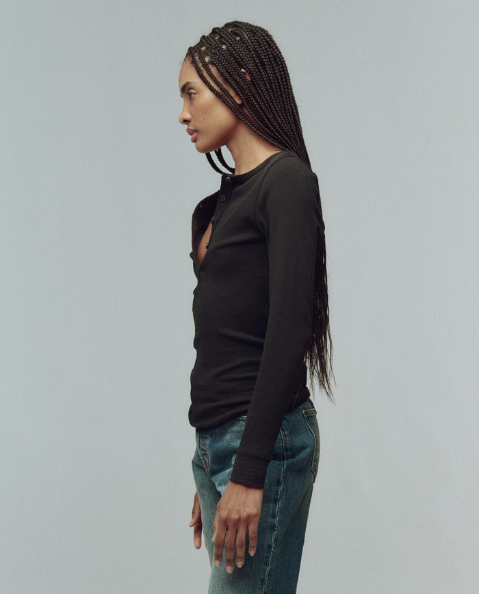 A person with long braided hair stands in profile, wearing THE GREAT. F25 BABY RIB's The Baby Rib Henley in Almost Black and blue jeans, set against a plain light background.