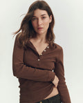 A woman stands against a plain background, wearing THE GREAT. PF26 RIB KNITS The Baby Rib Henley in Chocolate with button details and black pants, looking directly at the camera.