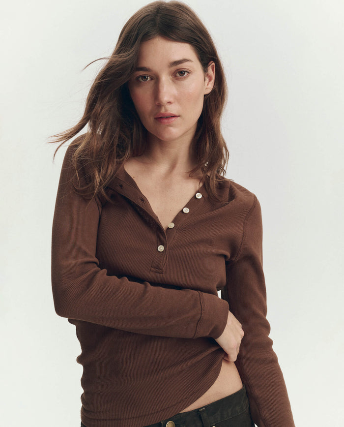 A woman stands against a plain background, wearing THE GREAT. PF26 RIB KNITS The Baby Rib Henley in Chocolate with button details and black pants, looking directly at the camera.
