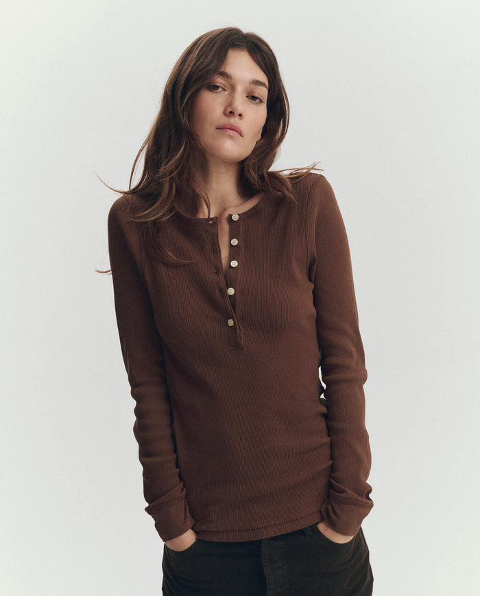 A woman with long brown hair wears THE GREAT. PF26 RIB KNITS The Baby Rib Henley in Chocolate and dark pants, standing against a plain light background.